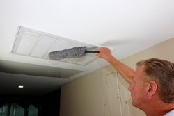 Rectangle white intake grid to a furnace being dusted with a microfiber gray dust wand by an adult man. Mature male dusting a rectangle ceiling HVAC intake grid of a home furnace to reduce allergens.