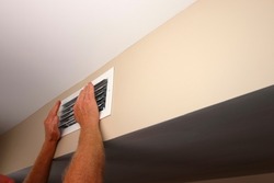 Feeling the air from a home air conditioning and heating HVAC vent on a wall are two hands in front of air duct grid. In a home near a ceiling is a rectangle white air vent with two hands in front of