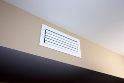 A white rectangle heating and cooling outflow air vent from a duct of an indoor HVAC system in a beige color wall near a ceiling. A small white rectangle outflow air duct vent inside a modern home.