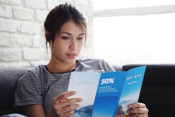 Young hispanic woman laying on couch at home, holding a travel flyer and planning next trip. Copy space on window.