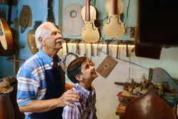 Small family business and traditions: old grandpa with grandson in lute maker shop. The senior artisan hugs the boy and shows him his handmade guitar and music instruments.