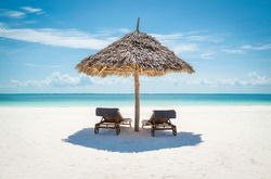 2 wooden sun loungers facing the tropical, turquoise blue Indian Ocean under a thatched umbrella on a white sandy Zanzibar beach
