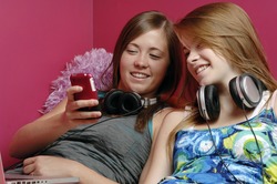 Two teenage girls use cellphone, laptop and a headset in their room.