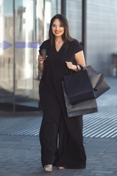 Street fashion week. Full body beautiful smiling overweight woman in total black outfit, wearing stylish jumpsuit, walking near shopping mall with many purchases in paper shopper bags.