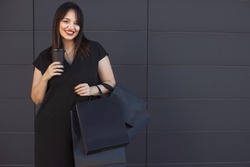 Street portrait of beautiful plus size happy woman with coffe cup and many purchases in black paper bags in buying at sale in shopping mall, standing on wall background. Black friday sale concept.