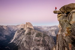 A male hiker standing on an overhanging rock at Glacier Point enjoying the breathtaking view towards famous Half Dome in beautiful twilight after sunset in summer, Yosemite National Park, California
