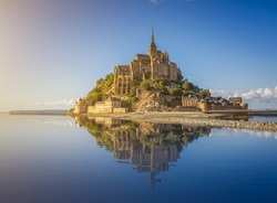Beautiful panoramic view of famous Le Mont Saint-Michel tidal island with deep blue water and clear reflections in golden evening light at sunset in summer, Normandy, northern France