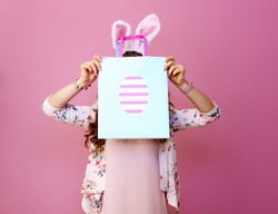 Festive bunny and eggs season. young woman in Easter bunny ears on pink background hide behind Easter shopping bag