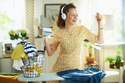 happy modern middle age woman in yellow dress with washed clothes basket ironing on ironing board while listening to the music with headphones at modern home in sunny day.