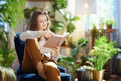 Green Home. relaxed trendy middle aged woman with long wavy hair with book in green pants and grey blouse in the modern living room in sunny day.