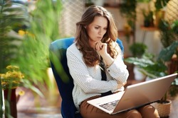 Green Home. pensive elegant 40 years old woman with long wavy hair in the modern living room in sunny day using laptop while sitting in a blue armchair.