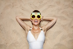 Warm sand treatment. Portrait of smiling woman in swimsuit and funny pineapple glasses laying on the sand
