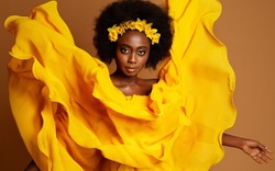 Dark Skinned Woman Fashion Portrait in Yellow Flying Dress. Beauty African Model with Flower Wreath in Black Afro Hairstyle. Dancer with fluttering Silk Fabric over Studio beige background