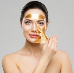 Beauty Woman peeling off Golden Facial Mask. Smooth Skin Model taking off Gold Purifying Face Film Mask over Gray. Women cleansing Cosmetics and Skincare Cosmetology