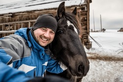 Happy man taking a selfie with black horse at winter ranch