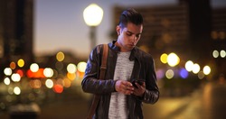 Millennial Hispanic man standing on city bridge at night looking at smartphone