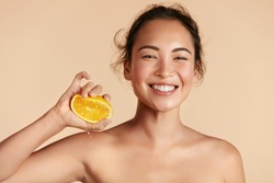 Beauty. Woman with radiant face skin squeezing orange in hand portrait. Beautiful smiling asian girl model with natural makeup, glowing facial skin and citrus fruit. Vitamin C cosmetics concept