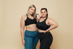 Body Positive. Plus Size Models Portrait. Confident Full-Figured Women In Sport Clothes Against Beige Background. Fitness For Active Lifestyle.