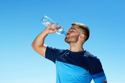 Man Drinking Water After Running. Portrait Of Handsome Athletic Male In Colorful Sportswear Resting After Fitness Workout, Drink Water From Bottle On Blue Sky Background. High Quality Image.
