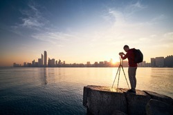 Photographer with camera on tripod photographing urban skyline at sunrise. Abu Dhabi, United Arab Emirates.