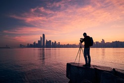 Silhouette of photographer with tripod. Young man photographing urban skyline. Abu Dhabi at dawn, United Arab Emirates