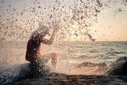 Unexpected wave of ocean splashing on man sitting on rock at sunset. 