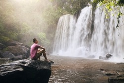 Man sitting on rock in front of high waterfall in mountains in tropical landscape in Camobodia.