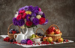 Autumn still life. A vase with asters, baskets of fruit, apples and plums and porcelain teaware on the table.
