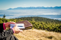 Outdoor Bible study during mountain hike. Female hands holding a Bible, empty notepad and pencil during outdoor devotion and Bible study in the mountains. Mountaintop outlook on High Tatras, Slovakia