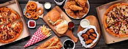 Table scene of assorted take out or delivery foods. Hamburgers, pizza, fried chicken and sides. Top down view on a dark wood banner background.