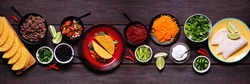 Taco bar table scene with a selection of ingredients. Overhead view on a dark wooden banner background. Mexican food buffet.