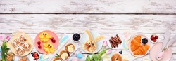 Easter breakfast or brunch bottom border. Top view on a white wood banner background. Bunny pancake, egg nests, chick fruit and an assortment of spring food items. Copy space.