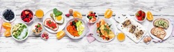 Healthy breakfast food table scene. Top down view over a white wood banner background. Omelette, nutritious bowl, toasts, granola bars, smoothie bowl, yogurts and fruits.