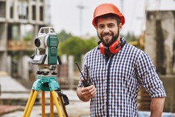 Surveyor equipment. Surveyor engineer in protective wear and red helmet using geodetic equipment, holding walkie talkie and smiling while standing at construction site