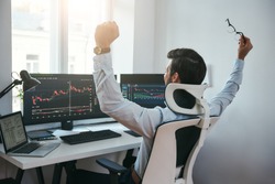 Yes! Back view of stock trader with raised hands looking at multiple computer screens with data and charts and feeling happy while sitting in modern office