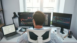 Trading stocks online. Back view of young businessman or trader working with graph and charts on computers at his modern office