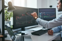 Forex market. Young trader is pointing at graphs on computer screen and analyzing data while working in his modern office.