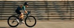 Website header of Side view of professional female cyclist in cycling garment and protective gear riding bicycle in city, rushing and passing buildings while training outdoors on a daytime