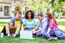 Young group of students with backpacks and books smiling, sitting on grass and using laptop. Campus as a background