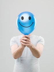 man hiding his face behing blue balloon with smiley face drawn on it, on grey background