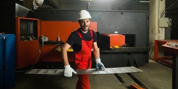 front view of a worker, looking happy, wearing red overalls, white protective helmet and gloves, standing, holding a sheet of metal plates in front of a red-painted heavy-duty machinery