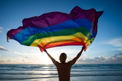 Silhouette of man holding a gay pride rainbow flag fluttering against the rising sun on an empty beach