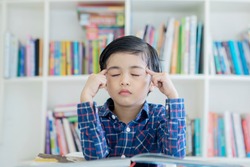 Image of a little boy thinking with eyes closed while learning in the library with bookshelf background