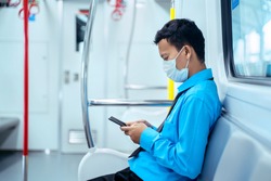 JAKARTA - Indonesia. May 12, 2020: Side view of male employee wearing a mask to prevention coronavirus while using a cellphone and sitting in the commuter train