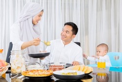 Muslim woman serving foods to her husband and daughter in dining room while having dinner during Eid Mubarak at home
