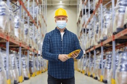 wholesale, logistic, people and export concept - male worker in safety helmet wearing face protective medical mask over warehouse background