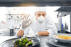 health, safety and pandemic concept - male chef cook wearing face protective mask or respirator for protection from virus disease with plate of soup and salad showing ok sign at restaurant kitchen