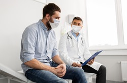 medicine, healthcare and pandemic concept - male doctor wearing face protective medical mask for protection from virus disease with clipboard and young man patient meeting at hospital