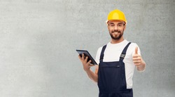 profession, construction and building concept - happy smiling male worker or builder in yellow helmet and overall with tablet pc computer showing thumbs up over grey concrete background