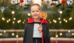 halloween, holiday and childhood concept - girl in dracula costume with black cape holding jack-o-lantern pumpkin party accessory over roof top background